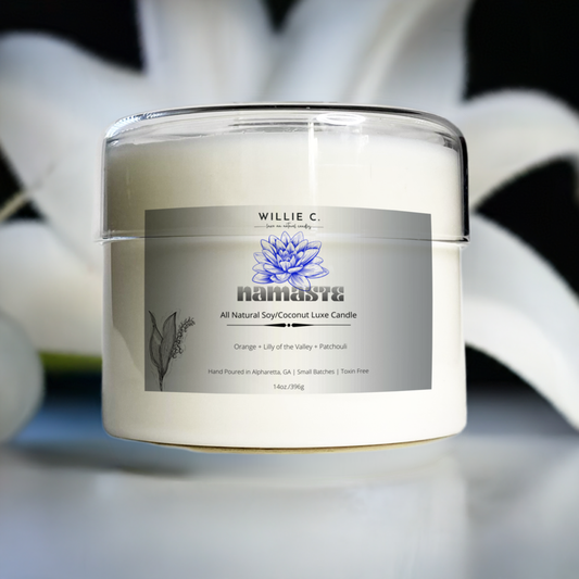 Glass candle container with glass lid sitting on a reflective surface with Namaste fragrance name. White lily behind container.